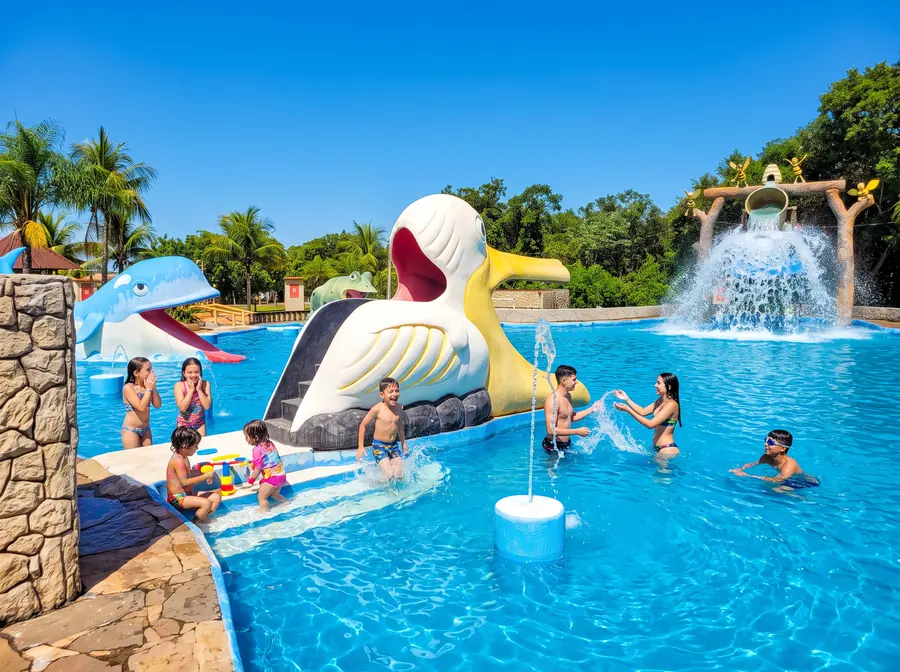 Baldão em forma de pelicano despejando água sobre a piscina, com famílias se divertindo
